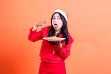 Yetişkin bir kadının Noel elbisesi, Noel Baba şapkası, baharatlı erişte yiyen sağ dil, ağızdan ağıza yelpaze, erişte dolu beyaz tabak, izole edilmiş turuncu arka plan.