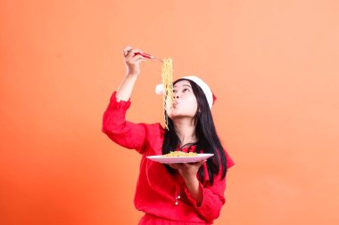 Noel elbisesi giyen, Noel Baba şapkası takan, erişteden zevk alan, kafasının üstünde erişte dolu bir çatal tutan ve turuncu arka planda izole edilmiş, erişteyle dolu beyaz tabak taşıyan kadınların ifadesi.