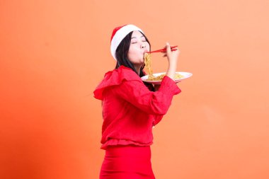 Endonezyalı kadın sol tarafta kapalı gözler erişteyi hissediyordu, yemek çubuklarını ağıza tutuyordu ve beyaz tabak erişteyle doluydu, kırmızı Noel bluzu giyiyordu, Noel Baba şapkası takıyordu, turuncu arka planda izole edilmişti.