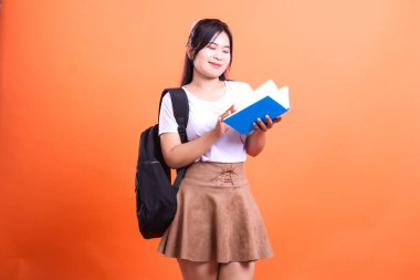 Siyah sırt çantalı bir kişi elinde kitap tutuyor ve okuyor. Arka plan katı turuncu bir renktir. Kişi beyaz gömlek ve kahverengi etek giyiyor..