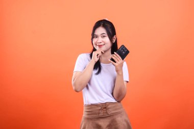 Genç bir kadın akıllı telefonuyla poz veriyor, kameraya bakıyor. O, beyaz bir tişört ve kahverengi bir etek giyiyor. Arkaplan sade turuncu renkte..