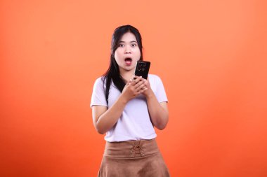 Şok olmuş bir yüz ifadesi olan genç bir kadın elinde bir akıllı telefon tutuyor. Turuncu bir arka planda izole edilmiş..