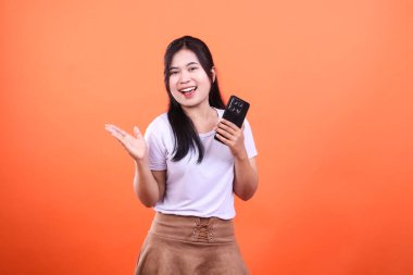 Genç bir kadın gülümsüyor ve sağ elinde siyah bir akıllı telefon tutuyor ve turuncu bir arka planda izole edilmiş sol eliyle bir şey sunuyor..