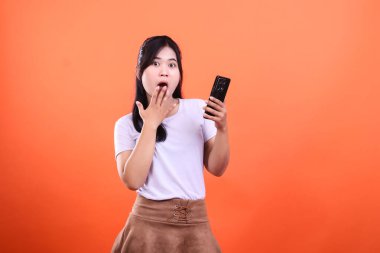 Genç bir kadın telefonunu turuncu bir arka planda tutarken şaşırmış görünüyor. Kadın beyaz bir gömlek ve kahverengi bir etek giyiyor. Eli açık bir ifadeyle ağzının yanında duruyor..