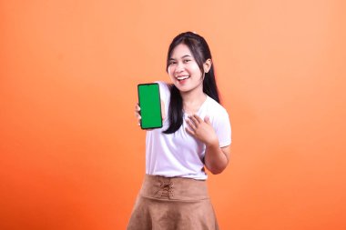 Beyaz tişörtlü ve kahverengi etekli bir kadın yeşil ekranlı bir cep telefonu sunar. Gülümsüyor ve telefonu kaldırıyor, turuncu arka planda tek başına..