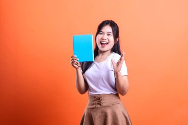 Bir genç elinde mavi bir kitap tutuyor ve kameraya gülümsüyor beyaz gömlek ve kahverengi etek giyiyor, turuncu arka planda izole edilmiş.