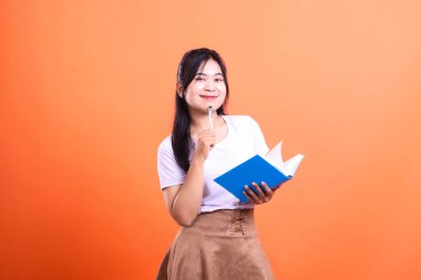 Genç bir kadın açık mavi bir kitap ve çenesinde bir kalem tutuyor, beyaz bir gömlek ve turuncu arka planda izole edilmiş kahverengi bir etek giyiyor.