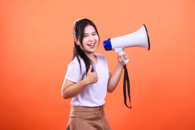 Beyaz gömlekli, kahverengi etekli ve turuncu arka planda duran kameraya gülümseyen genç bir kadın. Bir megafon tutuyor ve sol eliyle baş parmağını kaldırıyor.