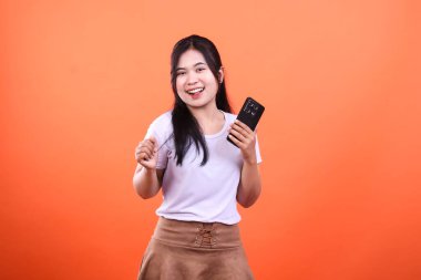 Beyaz tişörtlü ve kahverengi etekli bir kadın siyah bir cep telefonu tutuyor ve gülümsüyor..