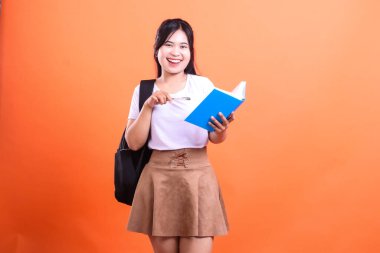 Elinde mavi bir kitap ve kalemle turuncu bir arka planın önünde duran genç bir kadın. O, beyaz bir tişört, kahverengi bir etek ve siyah bir sırt çantası giyiyor, gülümsüyor..