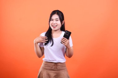 Uzun siyah saçlı, beyaz tişörtlü ve kahverengi etekli biri, siyah bir akıllı telefon tutuyor ve poz verirken gülümsüyor. Şüpheli turuncu bir arka planda izole edilmiş..