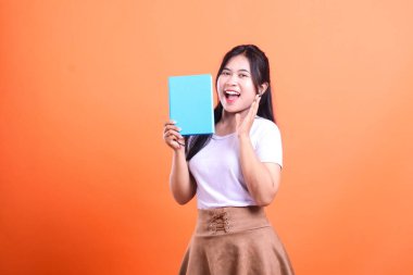 Bir kadın elinde sürpriz bir ifadeyle elinde mavi bir kitapla stüdyo ortamında poz veriyor. Beyaz bir gömleği ve kahverengi bir eteği var ve turuncu arka planda izole edilmiş.