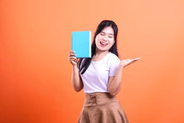 Mavi bir kitap tutan genç bir kadın, geniş bir gülümseme ve el hareketleriyle turuncu arka planda izole edilmiş bir şekilde el kol hareketi yapıyor..