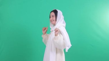 Sağında neşeli bir ifadeyle Asyalı tesettürlü Müslüman bir kadın, iki eli de dans ediyor, şarkının melodisini dinliyor, yeşil bir arka planda izole edilmiş, yeşil bir ekranda.