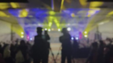 Oditoryumu dolduran bir grup bulanık insan sahne performansını izliyor. İki kişi bir video kamerayla kayıt yapıyor. Sahnede parlak bir ışık. Özet arkaplan, odak dışı