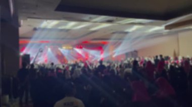 Oditoryumu dolduran bir grup bulanık insan sahne performansını izliyor. İki kişi bir video kamerayla kayıt yapıyor. Sahnede parlak bir ışık. Özet arkaplan, odak dışı