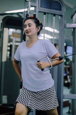 Asya 'lı bir kadın spor salonunda egzersiz makinesi kullanıyor. Sol kolu uzanmış, metal bir kabloyla bağlanmış, sağ kolu ise yan tarafında..