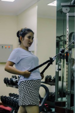 Asya 'lı bir kadın spor salonunda bir kablo makinesinde egzersiz yapıyor. Kadın makineye bağlı siyah bir ip tutuyor, kolları hafifçe bükülmüş ve ipi kendine doğru çekiyor..