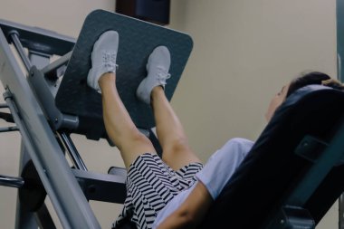 Bacak ütüleme makinesini kullanan kadınlar beyaz ayakkabı, şort ve beyaz gömlek giyiyorlar. Ayakları makinenin gri desenli ayak yatağında. Bacak pres makinesi gri metalden yapılır.