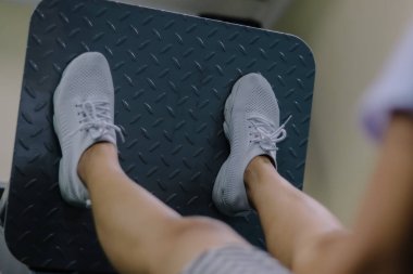 Bacak presi makinesinde bir kadının bacaklarını göstermek. İki ayağı da metal bir platformun ortasına yerleştirilmiş. Bağcıklı beyaz ayakkabılar ve kısa bir etek giyiyor.