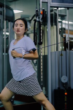 Asya 'lı bir kadın spor salonunda makine kullanıyor. Kabloya bağlı bir kablo tutuyor ve onu göğsüne doğru çekiyor..