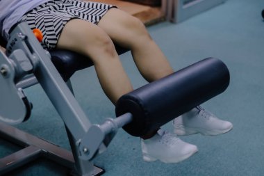 Bir kadının bacaklarını bacak uzatma makinesiyle kapatır, minderli bir sandalyeye oturur, bacaklarını makinenin bir parçası olan karanlık yastıklı bir çubuğun arkasına dayar.