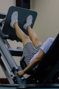 Kadın Asya spor salonunda bacak pres makinesi kullanıyor beyaz ayakkabılar ve kısa etek giyiyor ve bacaklarıyla makineye bastırıyor. Elleri makinenin saplarını tutuyor.