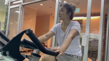 Spor salonunun içinde durgun bir bisiklette egzersiz yapan bir kadın görülüyor, elleri gidonu tutuyor. Beyaz bluz ve çizgili siyah beyaz kısa şort giyiyor. Odada cam duvarlar var.