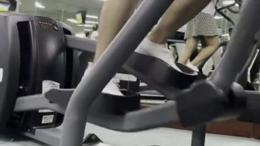 Spor salonunda egzersiz makineleri kullanan insanların yakın çekimi. Hareket edebilen ekipmanlar ve beyaz spor ayakkabıları üzerinde yoğunlaşıyoruz. Bir iç mekan görüntüsü.