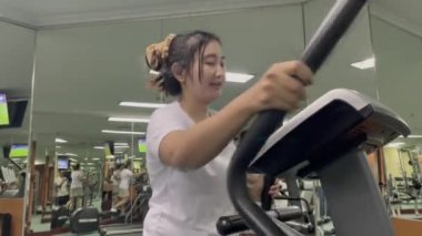 Beyaz tişörtlü bir kadın spor salonunda eliptik bir makine kullanırken görülüyor. Arka planda diğer insanlar ve egzersiz ekipmanları da görünür..
