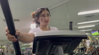 Bir kadın spor salonunda eliptik bir makine kullanıyor. Egzersiz yaparken saplarını tutuyor. Arka planda diğer ekipman ve insanlar görülebilir.