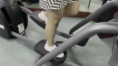 Bir kişi eliptik egzersiz makinesi kullanıyor. Beyaz ayakkabılar ve siyah beyaz bir elbise giyiyorlar. Makine yeşil halılı bir odada..