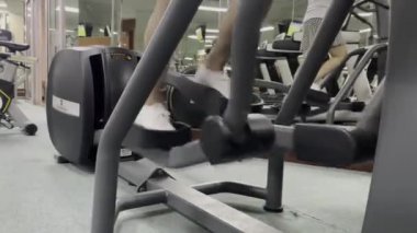 Spor salonunda bir kişi eliptik bir makine kullanıyor, beyaz ayakkabı giyen kişiyi kapatın. Arka planda başka insanlar ve ekipmanlar var..