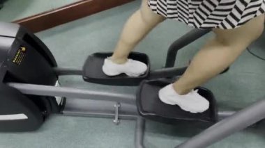 Eliptik bir makine üzerinde egzersiz yapan bir insan vücudu. Ayakları pedalların üzerinde ve egzersiz hareketleri görülebiliyor. Beyaz ayakkabılar ve kısa bir etek giyiyorlar..
