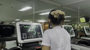 Saç tokası, beyaz gömlek ve kulaklıklı bir kadın spor salonunda aynalı bir koşu bandı kullanıyor. Arka planda diğer spor aletleri görülüyor. Koşu bandının dijital görüntüsü egzersiz verilerini gösterir.