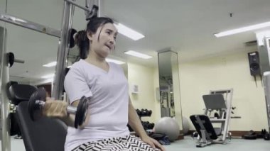 Beyaz tişörtlü ve desenli pantolonlu bir kadın spor salonunda çalışıyor. O, bir makinenin üzerinde otururken siyah bir dambıl kaldırıyor. Diğer spor aletleri görünür..