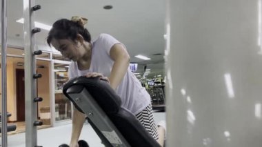 Bir kadın spor salonundaki yaslanmış bir bankta spor yaparken görülüyor. Arka planda spor aletleri ve televizyon ekranları var. Kadın beyaz bir gömlek giyiyor..