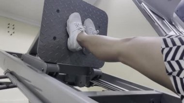 Spor salonunda bacak pres makinesi kullanan biri. Ayakları beyaz spor ayakkabılarla tabağa konur. Makine gri metaldir. Çizgili bir bluz giyiyor..