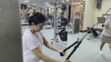Bir kadın spor salonunda idman yapmak için halat makinesi kullanıyor. Arka planda bir adam duruyor. Spor salonunda çeşitli egzersiz makineleri var. Sahne kapalı..
