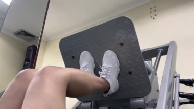 Bir kişi egzersiz için bacak baskı makinesi kullanıyor. Beyaz ayakkabılar ve desenli bir etek giyiyorlar. Bronz duvarlı bir spor salonundalar..