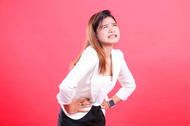 Beyaz ceketli ve siyah pantolonlu bir kadın iki eliyle karnını tutar ve kırmızı arka planda izole edilmiş bir acı ifadesi gösterir.