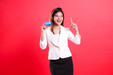 Beyaz takım elbiseli bir kadın kredi kartı tutuyor ve işaret parmağıyla işaret ediyor. Kırmızı arka planda izole bir şekilde gülümsüyor.