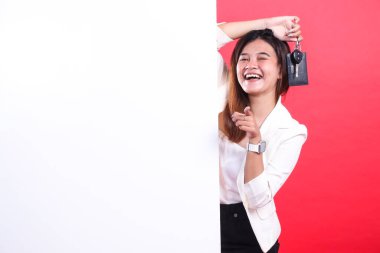 Gösterirken arabanın anahtarlarını tutan, beyaz bir bölümün arkasından bakan beyaz ceket ve kırmızı arka planda izole edilmiş siyah pantolon giyen bir kadın.