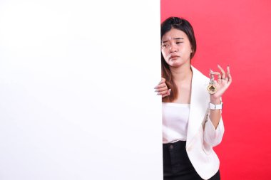 Uzun kahverengi saçlı bir kadın, üzerinde küçük bir ev olan bir anahtar tutuyor. Kısmen beyaz bir tahtanın arkasına gizlenmiş, kırmızı arka planda izole edilmiş.