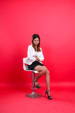 Beyaz takım elbiseli uzun boylu Asyalı bir kız, somurtuyor, kollarını kameraya kavuşturmuş, kırmızı arka planda izole edilmiş hidrolik sandalyede oturuyor.