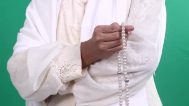 Bir el gümüş aksanlı inci dua boncuklarını tutuyor. El, yeşil arka planda ya da yeşil ekranda ince dantelli beyaz elbiseler giyiyor..