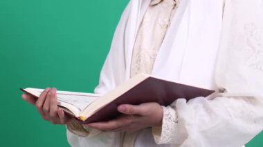 Beyaz cüppeli bir kişi iki eliyle süslenmiş kırmızı ve altın bir kitap tutuyor, yeşil arka planda ya da yeşil ekranda izole edilmiş..