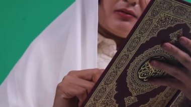 Beyaz başlıklı bir kadın elinde kırmızı ve altın desenli süslü bir Kuran tutuyor ve okuyor. yeşil arkaplanda ya da yeşil ekranda izole edilmiş