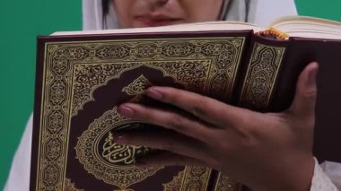 Bir kişi bir Kuran tutuyor ve onu okuyor, kapaktaki dekoratif detayları gösteriyor, ve yeşil arka plan veya yeşil ekrana izole edilmiş el ve kitaplara odaklanıyor.