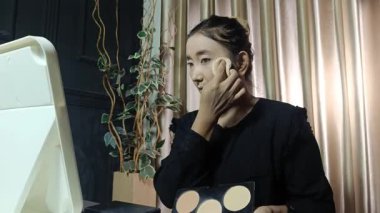 Genç bir Asyalı kadın yüzüne sünger kullanarak makyaj tozu sürüyor. O, aynaya bakıyor. Güzellik konsepti.